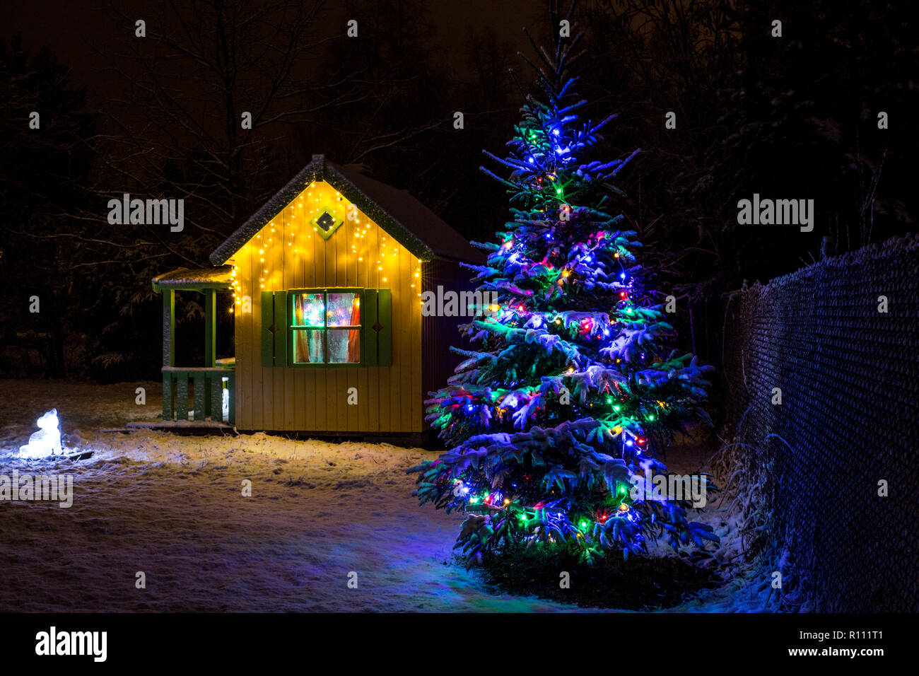 Wooden painted yellow private children`s play house in home garden, decorated with Christmas LED string lights outdoors in cold winder night. Decorate Stock Photo